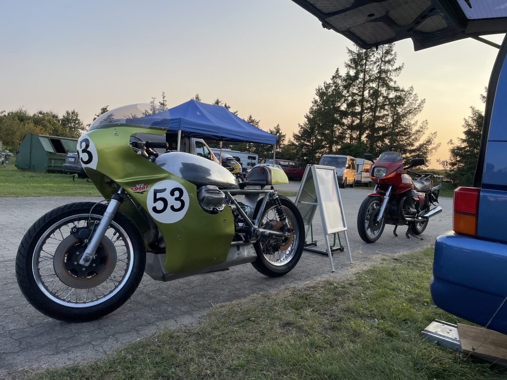 Parkeret Moto Guzzi V7 Sport med grøn fuldkåbe og aluminiumstank. På kåben står nummer 53 med sort tekst på hvid baggrund. Bagved holder Uffes røde Le Mans III. Billedet er fra Ring Djursland i forbindelse med næstsidste afdeling i DM i Road Racing.