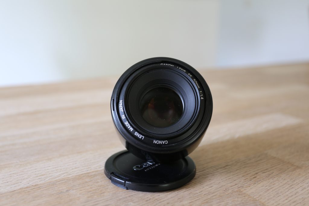 EF50mm f/1.4 USM, set forfra liggende på bord, med objektivdækslet under, så det vipper op i mod beskueren. Man kan se teksten på objektivets forside, samt støvpartikler på forreste linseelement.