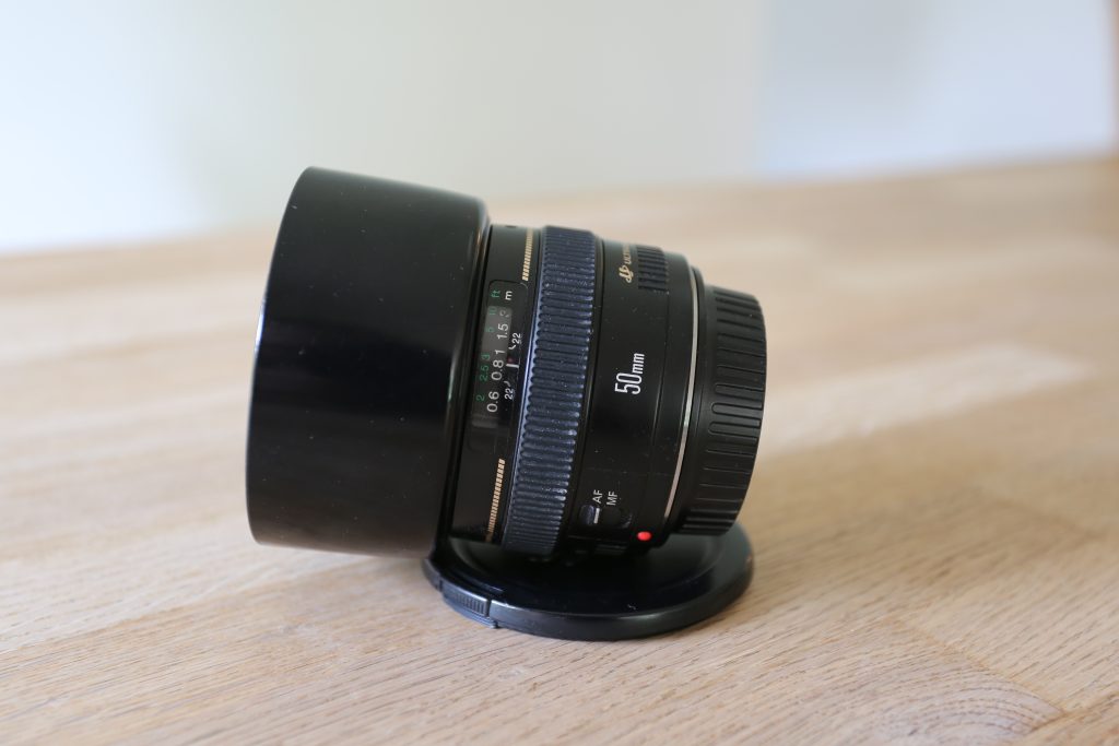 EF50mm f/1.4 USM, set fra siden liggende på bord, med objektivdækslet under. Man kan se  objektivets afstandsskala og fokusring. Objektivet har monteret modlysblænde (lens hood).