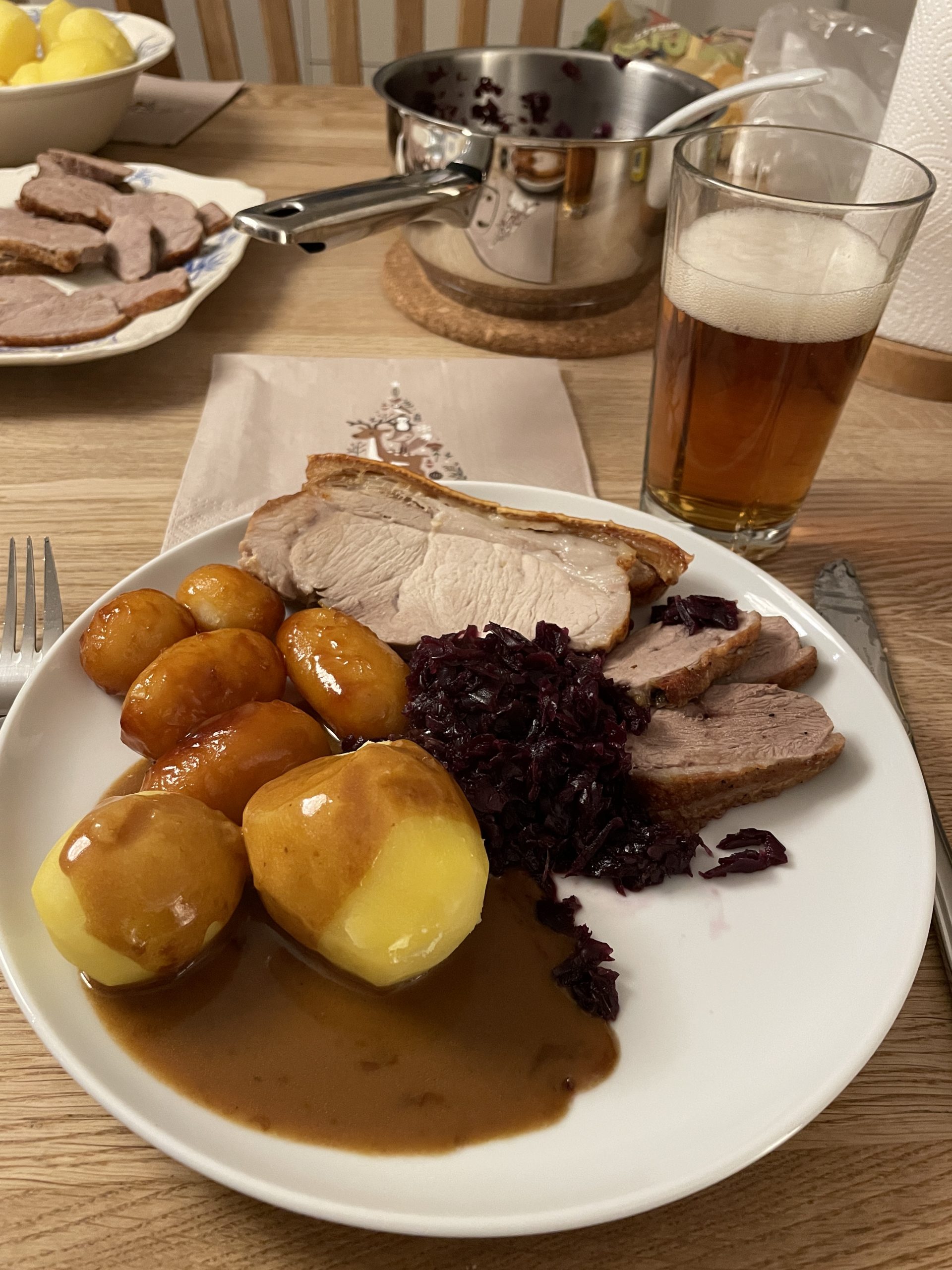 Tallerken med flæskesteg, stykker af stegt andebryst med røkål, hvide akartofler med brun sovs og brunede kasrtofler. Bag tallerkenen står er glas med øl.